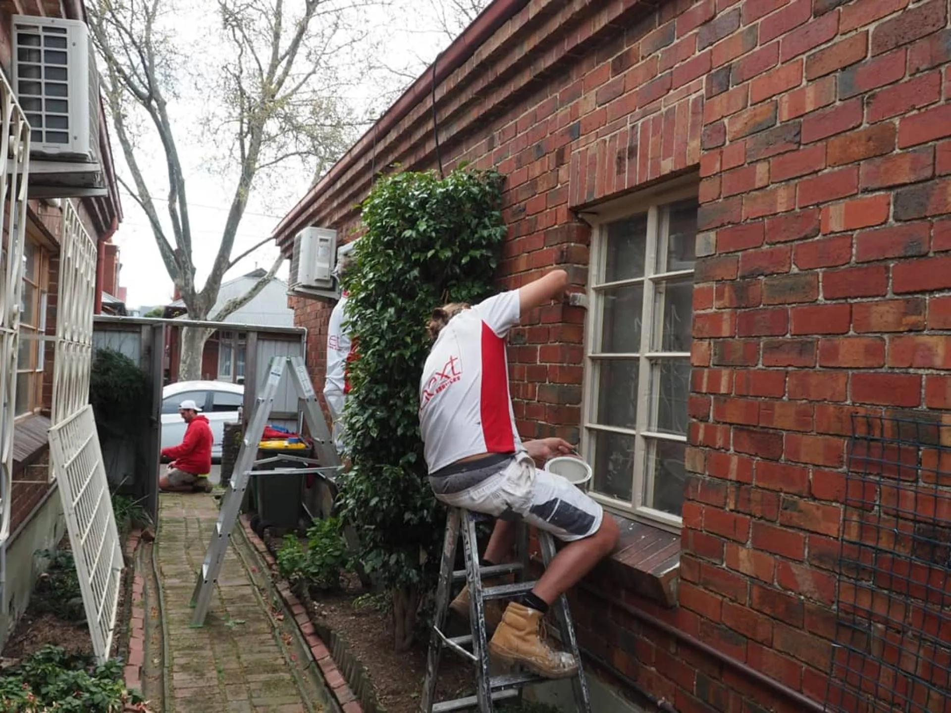 Two Next Painting painters in branded uniforms preparing timber window frames on a heritage red-brick Melbourne home