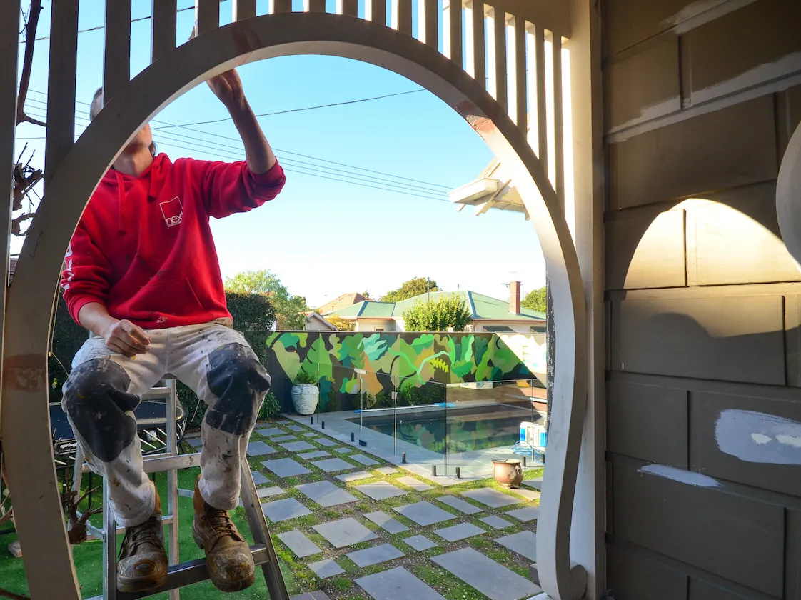 A Next Painting painter in a red branded hoodie on a ladder, painting an arched timber gate in a Prahran courtyard